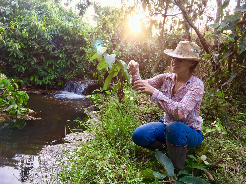 Dr. Marcia Macedo collecting a sample to measure greenhouse gases (methane and carbon dioxide) dissolved in water downstream of a reservoir in Mato Grosso, Brazil. Samples are collected in the field for later laboratory analysis at Woodwell Climate Research Center. 