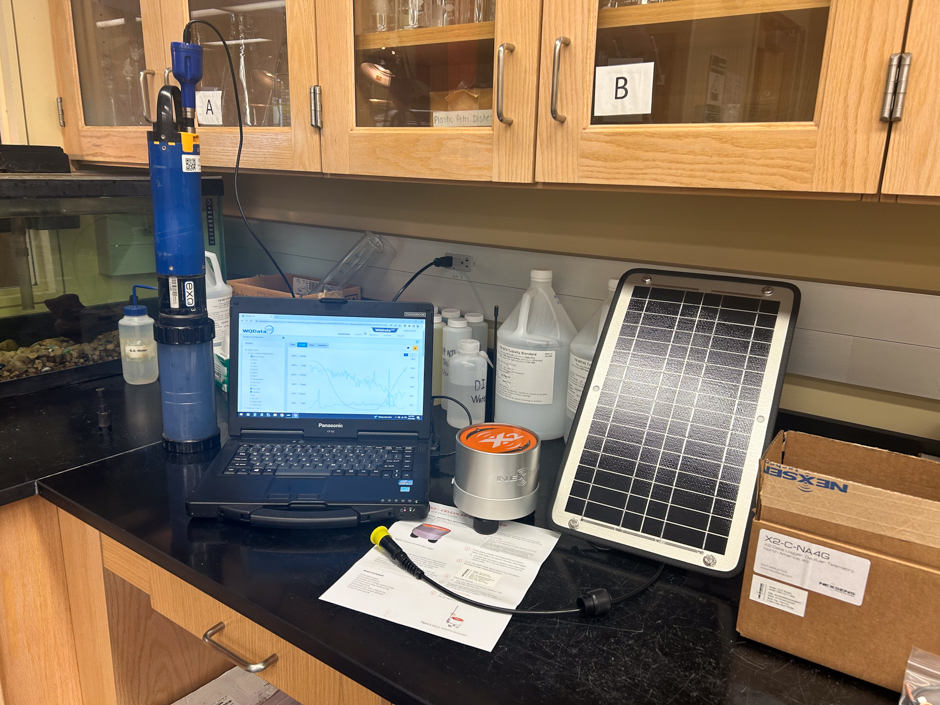The workbench at the NCWQR is set up for preparing an X2 logger for deployment with an EXO 3 sonde, both of which will be powered by the solar panel in the picture. The WQDataLive portal is open on the laptop with graphs of current data from a different nutrient sampling location.