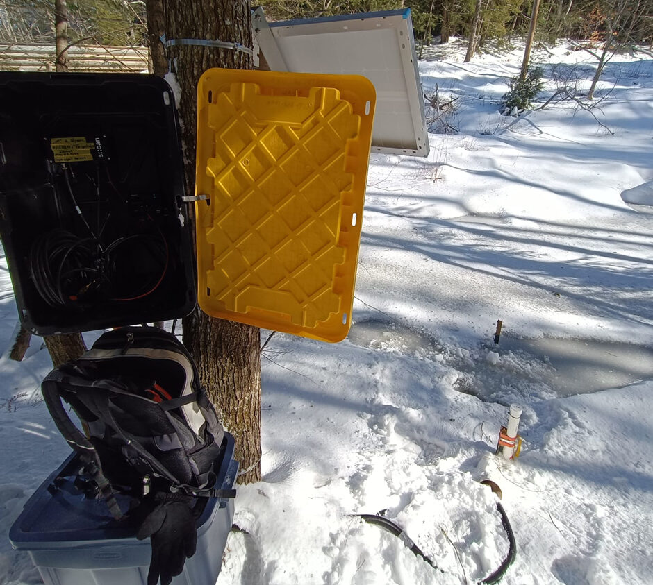 Datalogger and battery charger housed in a box mounted on a tree. Below it, a second box contains a large battery designed to support low-light conditions, such as during winter. Behind the main box, a solar panel supplies energy. Note the frozen river surface, where a PVC pipe holds a HOBO U20 sensor.